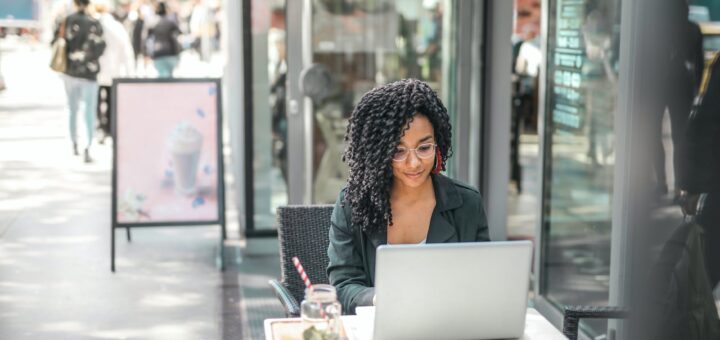 ethnic young woman using laptop while having tasty beverage in modern street cafe